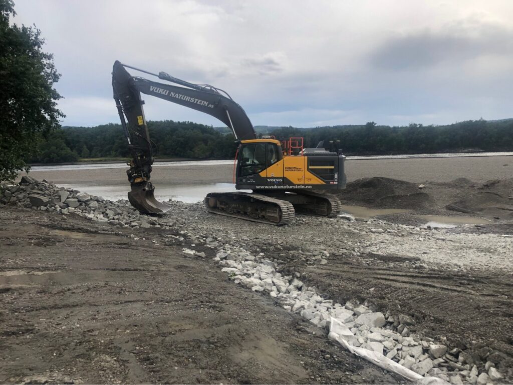 Foto av parkert gravemaskin i skråning ned mot vann